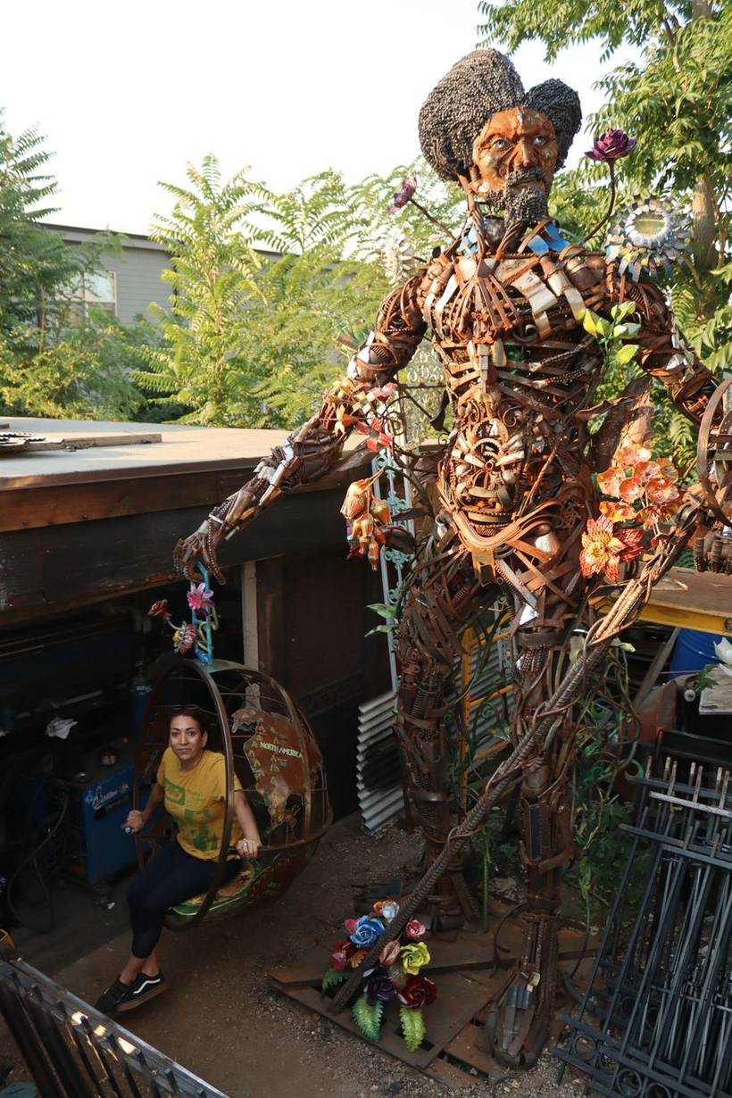 Welded scrap-iron figure with painted metal flowers standing in the Superior Iron Works yard, a globe chair at left and stacked iron fence panels at right